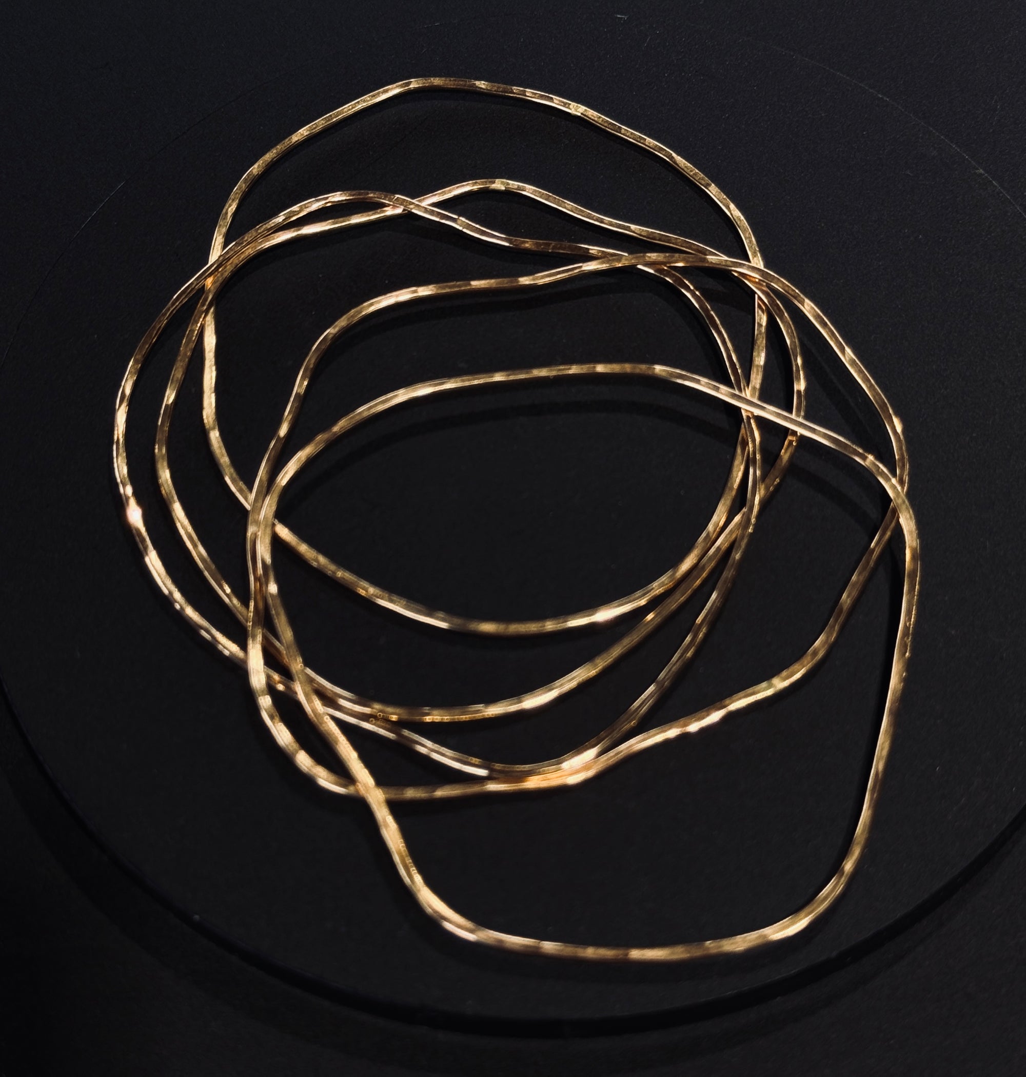 A group of five gold metal bangle bracelets with a hammered or textured finish, displayed on a dark surface.