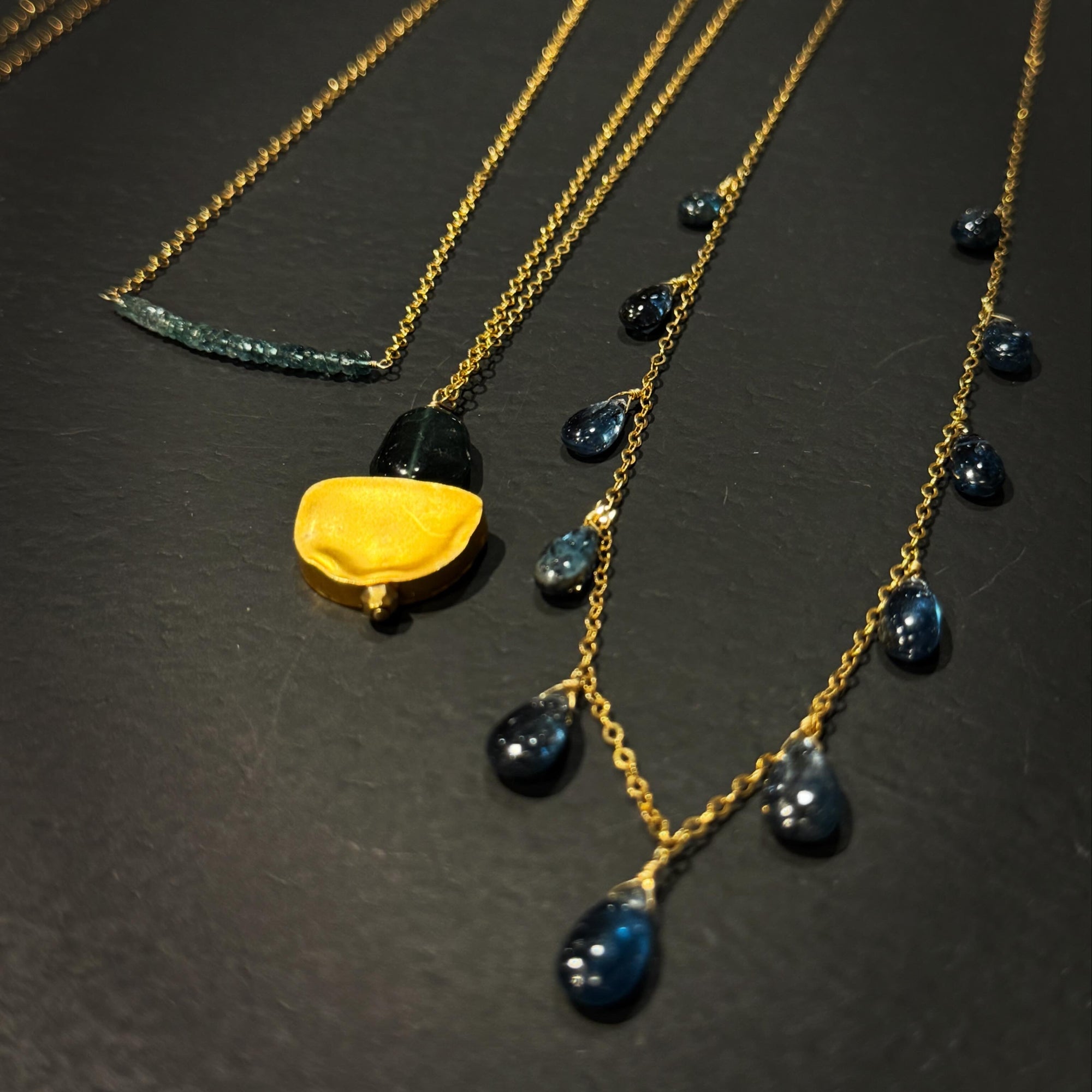 Gold-tone layered necklaces displayed on a dark textured surface. One necklace features a bright gold textured crescent moon bead and dark teal stone, while another has small dark blue teardrop gemstones, and a third has a curved dark blue green stone accent.